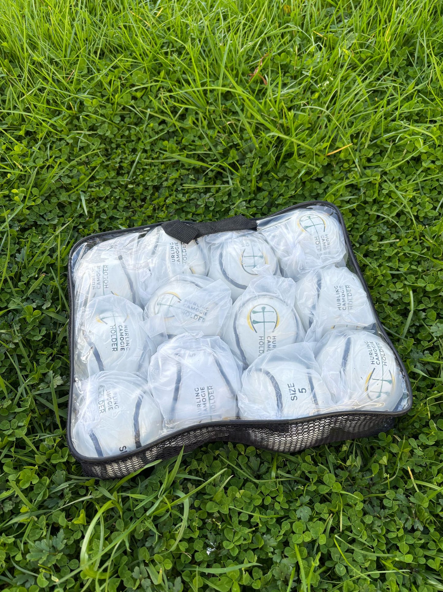 A white sliotar, which is a type of field hockey ball, lying on green grass next to a red and black hurling stick with a logo on the grip.