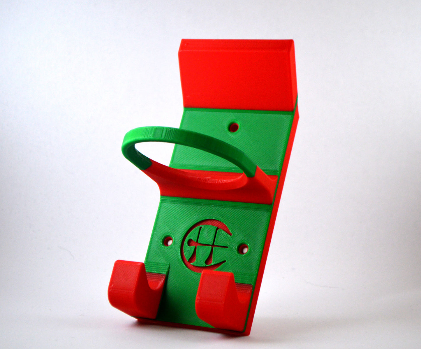 A red and green hurling holder with a logo cut into the design, featuring a helmet hook and a hurley hook and a sliotar holder, placed against a grey background.