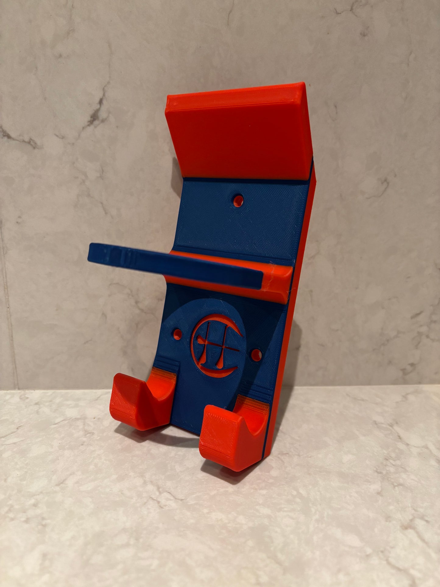 A blue and red hurling holder with a logo cut into the design, featuring a helmet hook and a hurley hook and a sliotar holder, placed against a grey background.