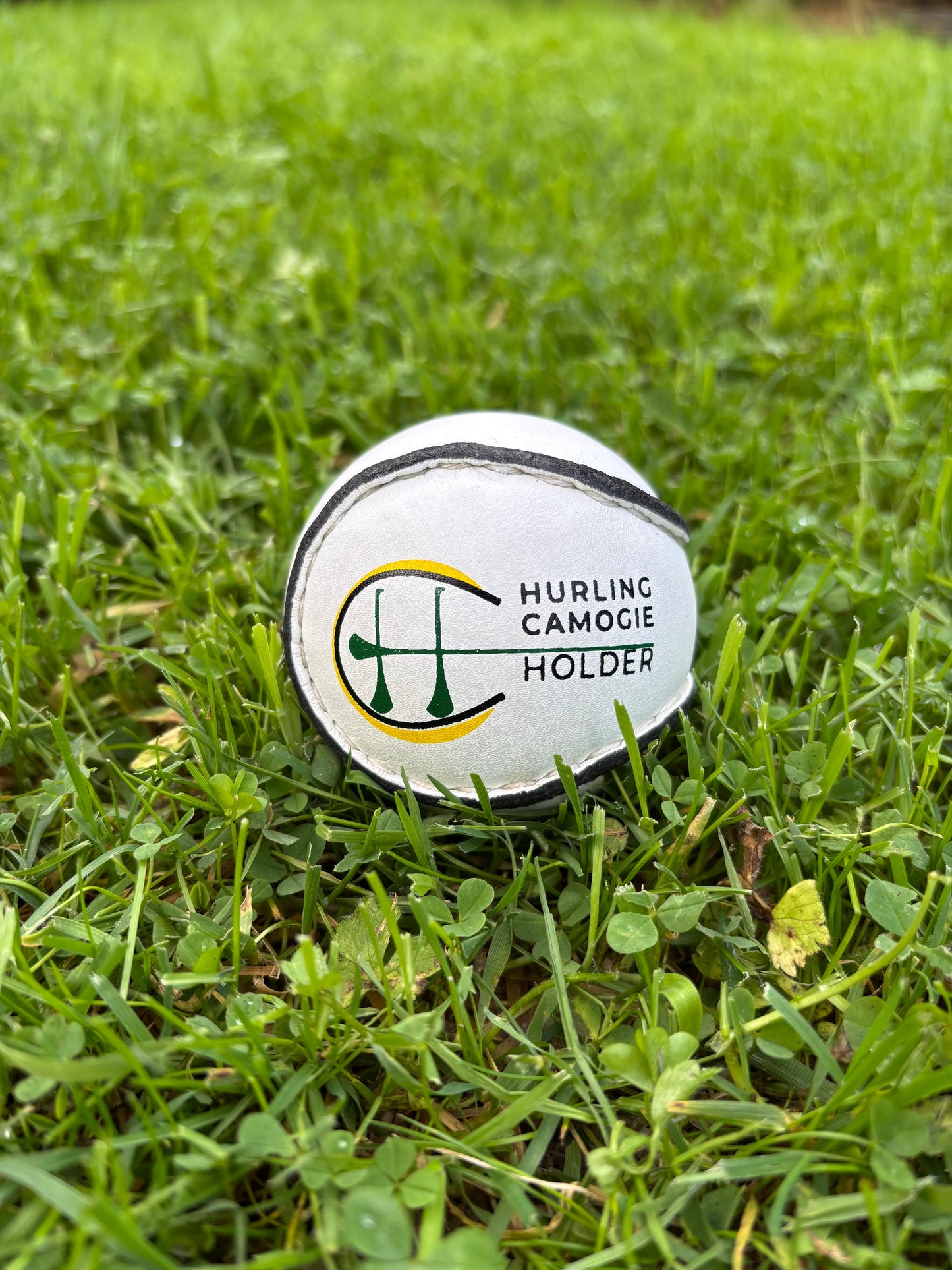 A white sliotar, which is a type of field hockey ball, lying on green grass next to a red and black hurling stick with a logo on the grip.