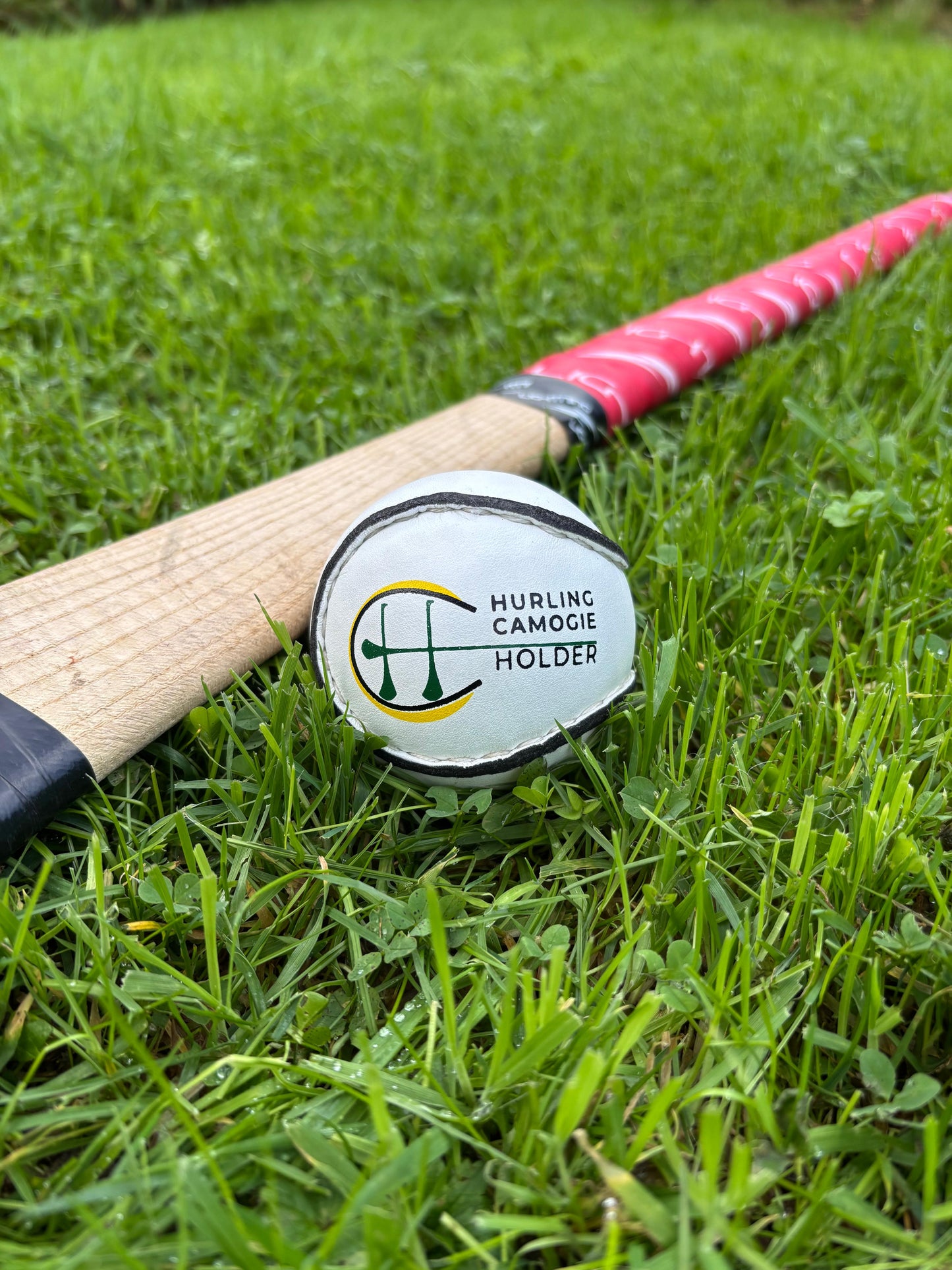 A white sliotar, which is a type of field hockey ball, lying on green grass next to a red and black hurling stick with a logo on the grip.