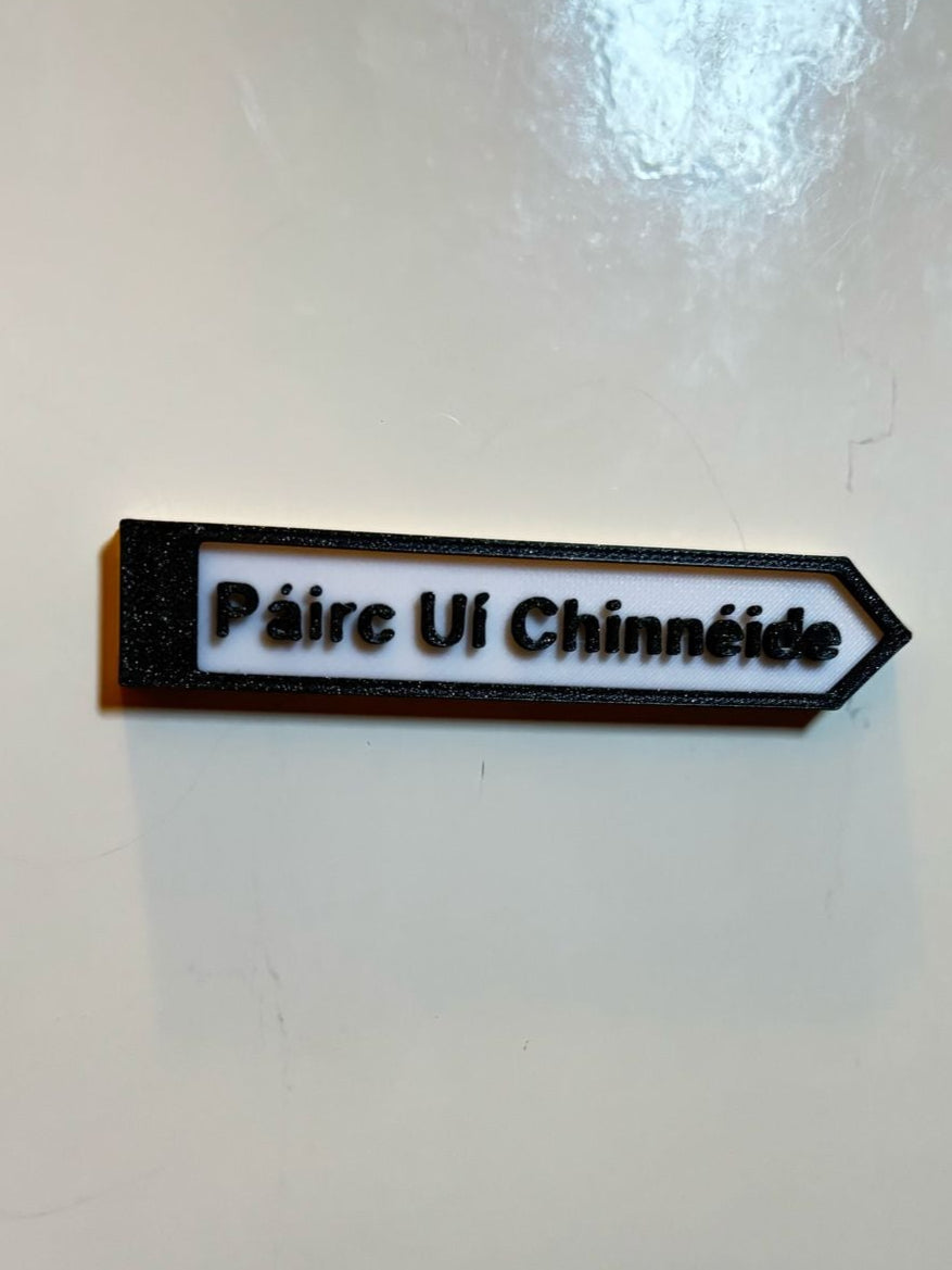 A black and white rectangular fridge magnet with white text 'pairc ui chinneide' on a white wall, representing a miniature size signpost.