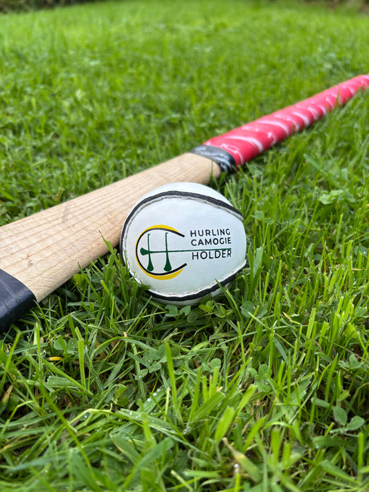 A white sliotar, which is a type of field hockey ball, lying on green grass next to a red and black hurling stick with a logo on the grip.